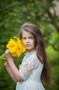 Güzel beyaz elbiseli şık bir kız yeşil yapraklı sarı karahindiba çiçekleriyle poz veriyor.
