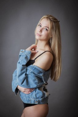 Siyah iç çamaşırlı ve kot ceketli ince bir kız koyu bir arka planda poz veriyor.