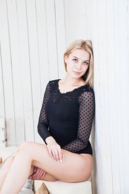 Siyah iç çamaşırlı, uzun sarı saçlı ince bir kız beyaz bir duvara poz veriyor.