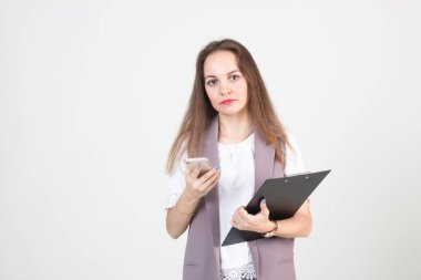 İş dünyasındaki genç ve güzel bir kadın. Katı giysiler. Elinde telefonla beyaz bir duvara poz veriyor.