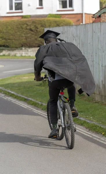 Andover, Hampshire, England, UK. May 2019. A rear view of a university student wearing cap and gown riding a bicycle.