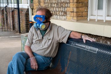 Hampshire, İngiltere, İngiltere. 2020 yılı. İngiltere 'de Covid-19 salgını sırasında tren istasyonunda oturan filtre ve gözlüklü bir maske takan adam..