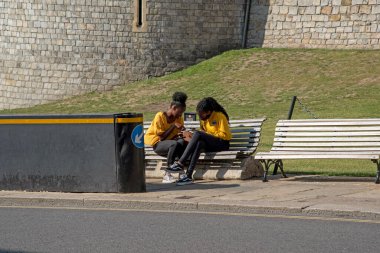 Windsor, Berkshire, İngiltere, İngiltere. 2020 yılı. Windsor Kalesi 'nin dışındaki ahşap bankta oturan turistler kaldırım kenarındaki güvenlik bariyerine yakın oturuyorlar..