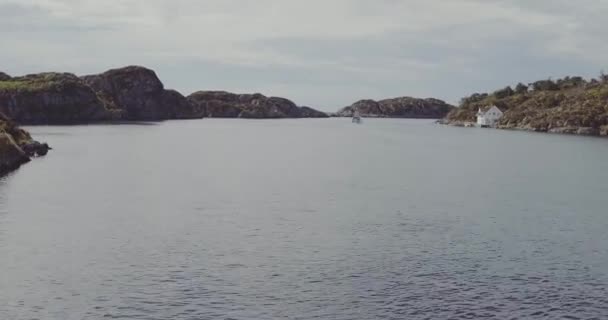 飞行在峡湾波纹水与遥远的小船反对岩石银行和单独的白色房子在山脚在灰色的天空之下 — 图库视频影像