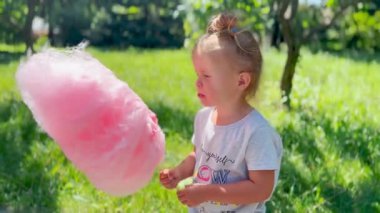 Küçük kız dışarıda pembe pamuk şekerin tadını çıkarıyor. Tatlı ikramların ve yaz kutlamalarının sembolü.