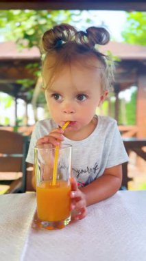 İki poposu olan tatlı küçük kız pipetle portakal suyu içiyor. Yaz günü, sağlıklı yaşam tarzı ve neşeli ruh hali.