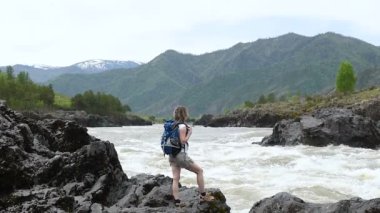 Bir kız bir sırt çantası ile bir dağ nehir kıyısında bir kayanın üzerinde duruyor. Ayaklarını bir dere köpüren.