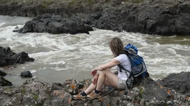 バックパックを持つ少女は 岩の上の山の川の銀行で置かれます ストリームのバブル彼女の足の下 — ストック動画