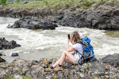 Bir kayanın üzerinde bir dağ nehir kıyısında bir sırt çantası ile bir kız oturuyor. Ayaklarını bir dere köpüren.