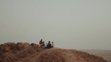 Gün batımında bir dağın tepesinde dinlenen bir grup insan, şenlik ateşinin yanında oturmuş, eğleniyorlar - arkadaşlık, gençlik, 4k