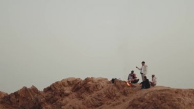 Gün batımında bir dağın tepesinde dinlenen bir grup insan, şenlik ateşinin yanında oturmuş, eğleniyorlar - arkadaşlık, gençlik, 4k