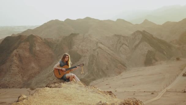 Mujer tocando la guitarra y cantando en el desierto en paisajes al atardecer, montañas del desierto de fondo, 4k — Vídeo de stock