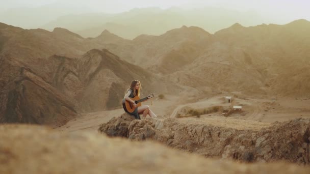 Kvinna som spelar gitarr och sjunger i öknen i solnedgången landskap, öken berg bakgrund, 4k — Stockvideo