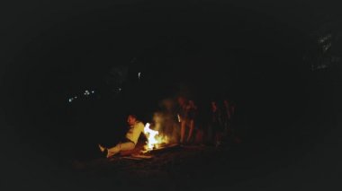 Bir grup arkadaş, gece deniz kenarında kamp ateşinin yanında eğleniyorlar.