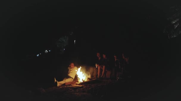 Grupo de amigos divirtiéndose cerca de la fogata en la playa cerca del mar por la noche, 4k — Vídeo de stock