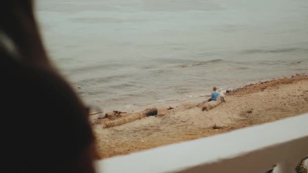 Samotna kobieta pije kawę lub herbatę rano na balkonie z niebieskim widokiem na morze, dzieci bawią się na plaży w pobliżu morza, 4k — Wideo stockowe