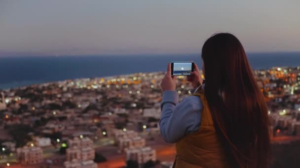 Mujer tomar una foto del mar por teléfono inteligente en la noche. Mujer permanecer en la cima de la montaña y mirando en Horizonte, 4k — Vídeo de stock