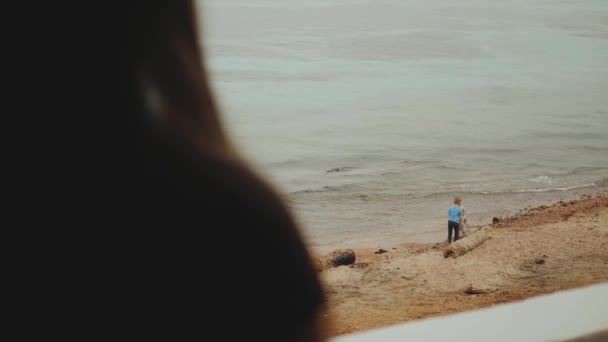 Samotna kobieta pije kawę lub herbatę rano na balkonie z niebieskim widokiem na morze, dzieci bawią się na plaży w pobliżu morza, 4k — Wideo stockowe