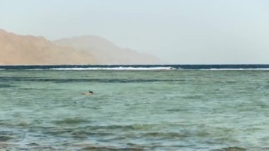 Kızıl denizde şnorkelle yüzen adam, güzel mavi deniz manzarası ve berrak gökyüzü, denizdeki dalgalar ve ufuktaki dağlar, Mısır, Dahab, 4k