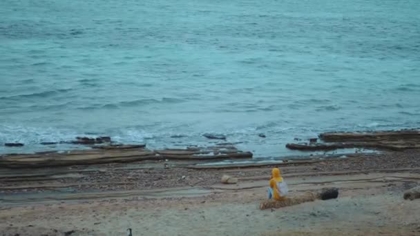 Mooie vrouw zit alleen op stenig strand in de buurt van de zee, de golven breken op de kust, Egypte Sinai, 4k — Stockvideo