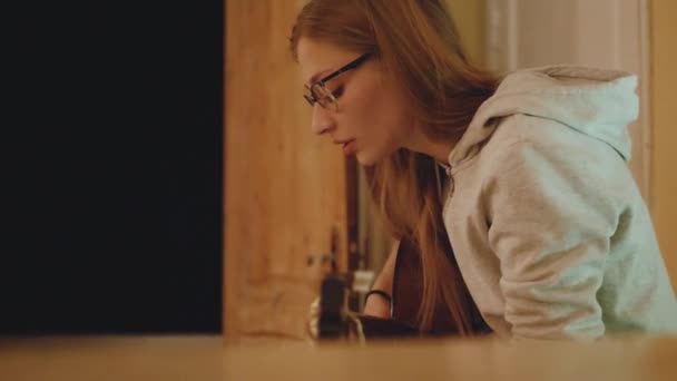 A mulher loira de óculos toca guitarra e canta na cadeira em apartamento acolhedor. Uma mulher cria música dentro de casa, 4k — Vídeo de Stock