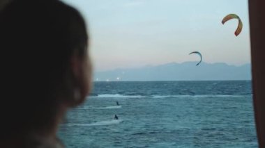 Kadın şarap içiyor ve bak insanlar Dahab Mısır 'da temiz suda nasıl sörf yapıyor. Arka planda dağlarla mavi denizi keşfetmek ve insanlar uçurtma sörfü yapıyor, 4k