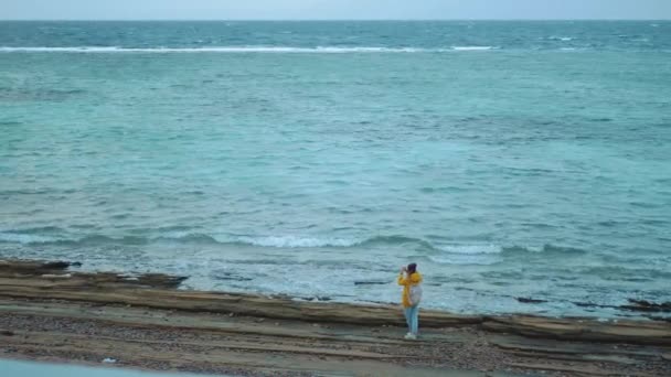 Jolie femme marchant et prenant des photos sur la plage de rivage pierreux près de la mer, les vagues se brisent sur le rivage, temps froid, Egypte Sinaï, 4k — Video