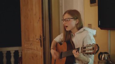 Gözlüklü sarışın kadın gitar çalıyor ve rahat bir dairede sandalyede şarkı söylüyor. Bir kadın içeride müzik yaratır, 4k