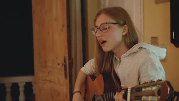 A mulher loira de óculos toca guitarra e canta na cadeira em apartamento acolhedor. Uma mulher cria música dentro de casa, 4k — Vídeo de Stock