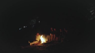 Bir grup arkadaş, gece boyunca deniz kenarında kamp ateşinin yanında eğleniyorlar.