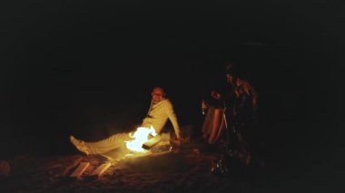 Bir grup arkadaş, gece boyunca deniz kenarında kamp ateşinin yanında eğleniyorlar.