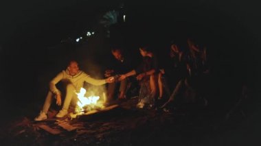 Bir grup arkadaş, gece boyunca deniz kenarında kamp ateşinin yanında eğleniyorlar.