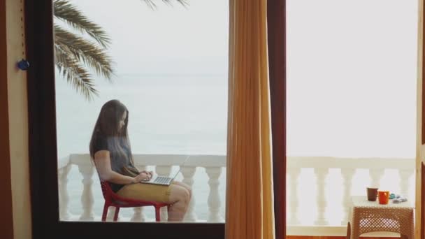 Студентка или фрилансер, работающая на ноутбуке, женщина, сидящая на балконе с видом на пальмы и море, полная. — стоковое видео