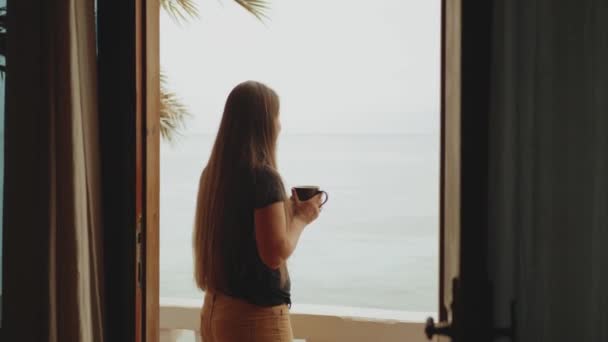 Μόνος γυναίκα πίνει καφέ ή τσάι το πρωί στο μπαλκόνι με μπλε θέα στη θάλασσα, άποψη από το παράθυρο, full hd — Αρχείο Βίντεο