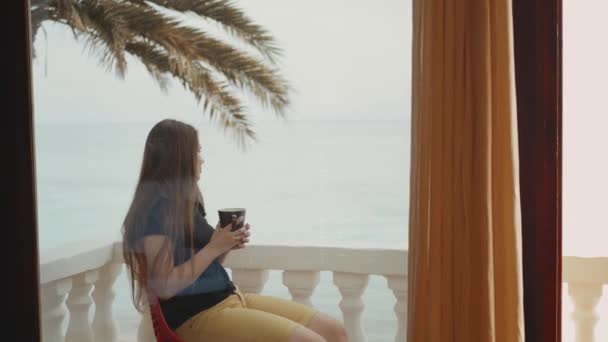 Μόνος γυναίκα πίνει καφέ ή τσάι το πρωί στο μπαλκόνι με μπλε θέα στη θάλασσα, άποψη από το παράθυρο, full hd — Αρχείο Βίντεο