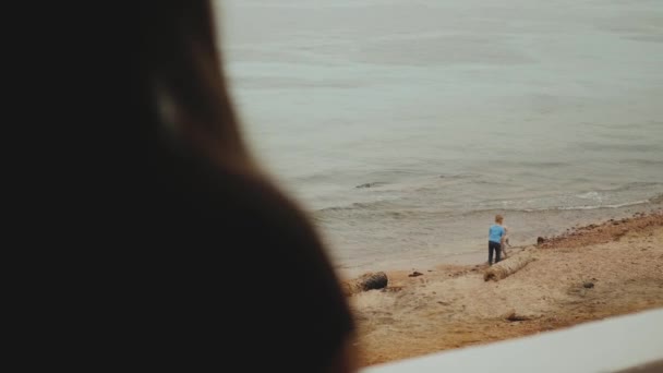 Sozinha que fica na varanda e para a vista azul do mar, as crianças brincam na praia perto do mar, hd completo — Vídeo de Stock