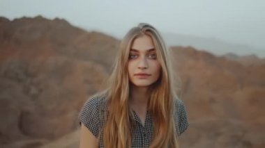 Elbiseli genç sarışın kadın dağın tepesinde yürüyor ve etrafında dönüyor. Gün batımlı dağ manzarası, tam HD