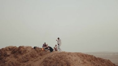 Gün batımında bir dağın tepesinde dinlenen bir grup insan, şenlik ateşinin yanında oturmuş, zamanlarının tadını çıkarıyorlar - arkadaşlık, gençlik, tam kapasite.