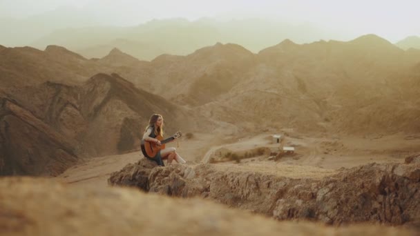 Mulher tocando guitarra e cantando no deserto em paisagens do pôr do sol, deserto montanhas fundo, hd completo — Vídeo de Stock