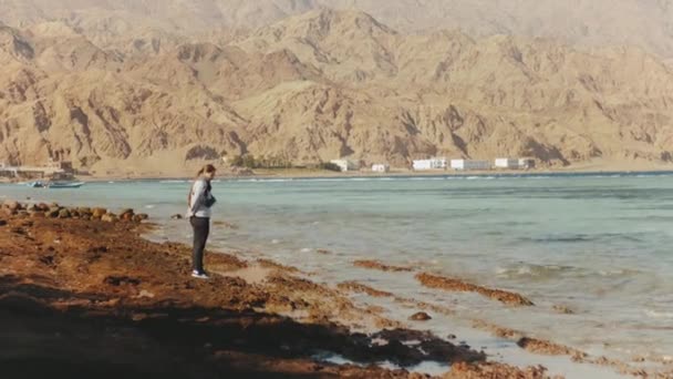 Mujer bonita caminando sola en la playa costera pedregosa cerca del mar, las olas se rompen en la orilla, Egipto Sinaí montaña en el fondo, hd completo — Vídeos de Stock