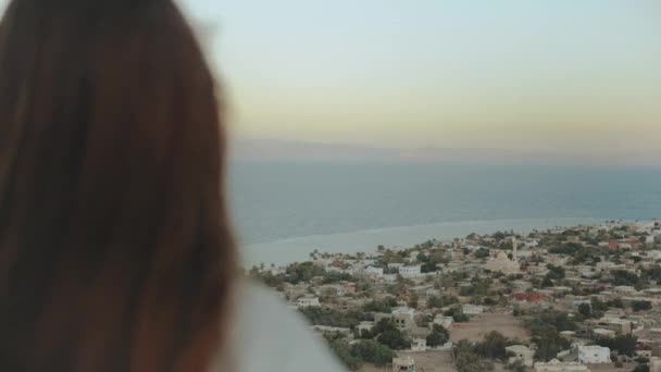 Вид на синее море и маленькую деревню со спины женщины. Девочка остается на вершине горы и смотрит на нее, полная сил. — стоковое видео