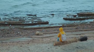 Sarı ceketli güzel bir kadın deniz kıyısındaki taşlı sahilde tek başına oturuyor, dalgalar kıyıya vuruyor, Mısır Sina, tam kapasite