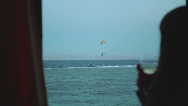 Zbliżenie kobiety pije wino i zobacz jak ludzie kitesurfują wieczorem na czerwonym morzu Egiptu. Punkt widzenia z okna, dwóch mężczyzn kitesurfing na horyzoncie, full hd — Wideo stockowe