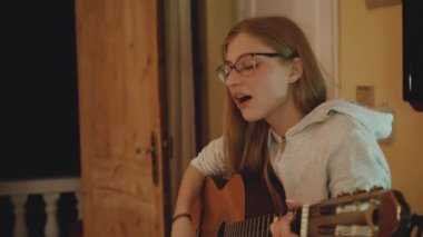 Gözlüklü sarışın kadın gitar çalar ve şarkı söyler, rahat bir dairede sandalyede oturur. Bir kadın kapalı mekanda duygusal müzik yaratır.