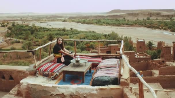 Hübsche Frau sitzt auf einer Dachterrasse mit einem schönen Blick auf die Ruinen einer verlassenen Stadt Die berühmte Ait Benhaddou Kasbah, Altstadt, Schöne touristische Orte Marokkos, 4k — Stockvideo