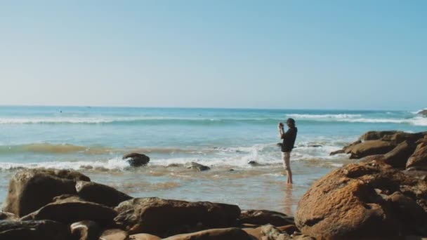 Homem tirando fotos no smartphone na praia, o oceano no fundo, costa pedregosa, turista em Marrocos, 4k — Vídeo de Stock