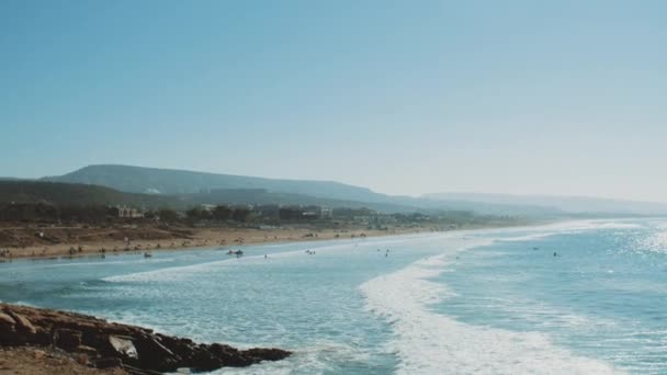 Surfing i Atlanten, Marocko, många människor på stranden, stora vågor och klar himmel och berg vid horisonten, Taghazout byn, 4k — Stockvideo