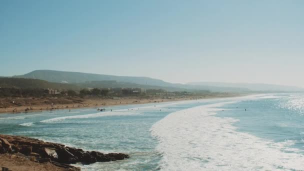 Surfing i Atlanten, Marocko, många människor på stranden, stora vågor och klar himmel och berg vid horisonten, Taghazout byn, 4k — Stockvideo