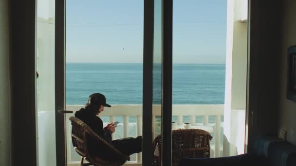 Jovem desfrutando de vista para o mar a partir do terraço. Viajante masculino olhando para o telefone na varanda com belo oceano Taghazout Marrocos, 4k — Vídeo de Stock