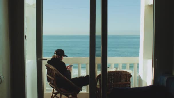 Giovane uomo godendo vista sull'oceano dalla terrazza. Viaggiatore maschile guardando il telefono sul balcone con bellissimo oceano Taghazout Marocco, 4k — Video Stock